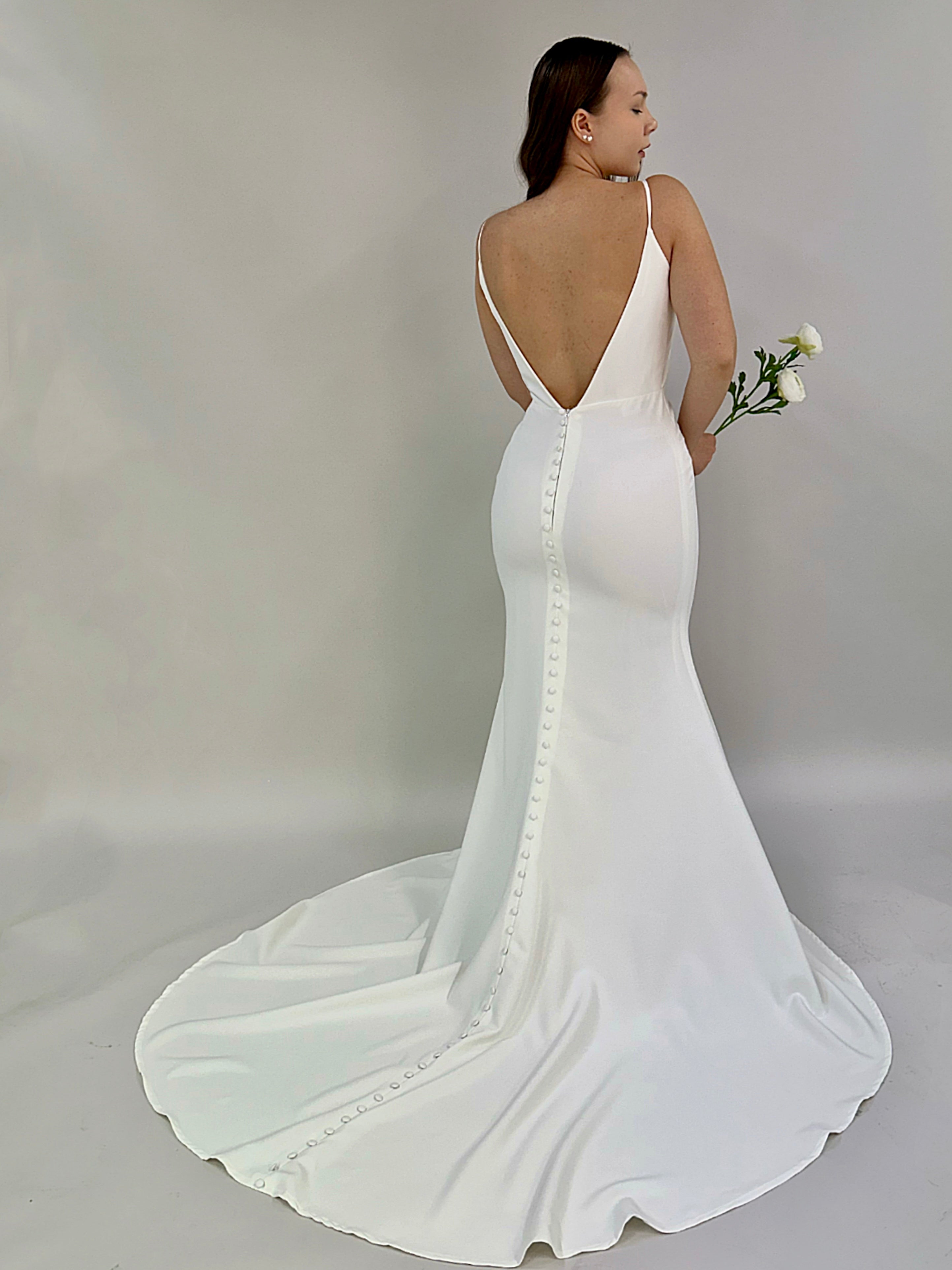 Minimalistinen häämekko ohuilla olkaimilla, syvään uurrettu V-selkä ja napitus selästä laahukseen.