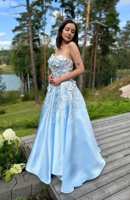 Vaaleansininen satiininen vanhojentanssimekko olkaimettomalla mallilla, koristeltu kukkapitsillä.